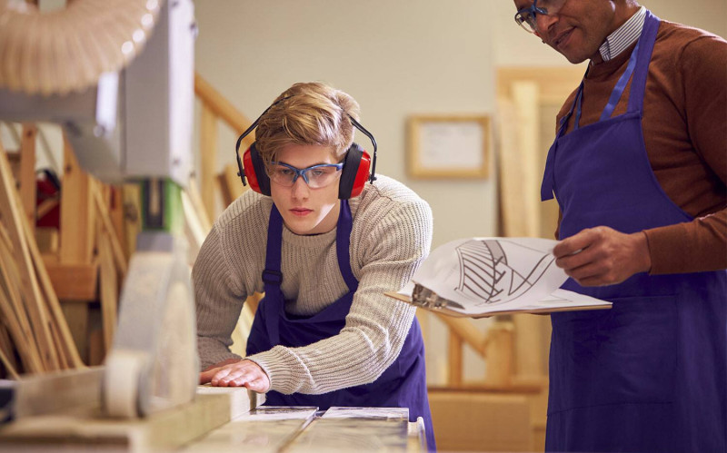 A teacher instructs a high school student in using woodworking equipment.